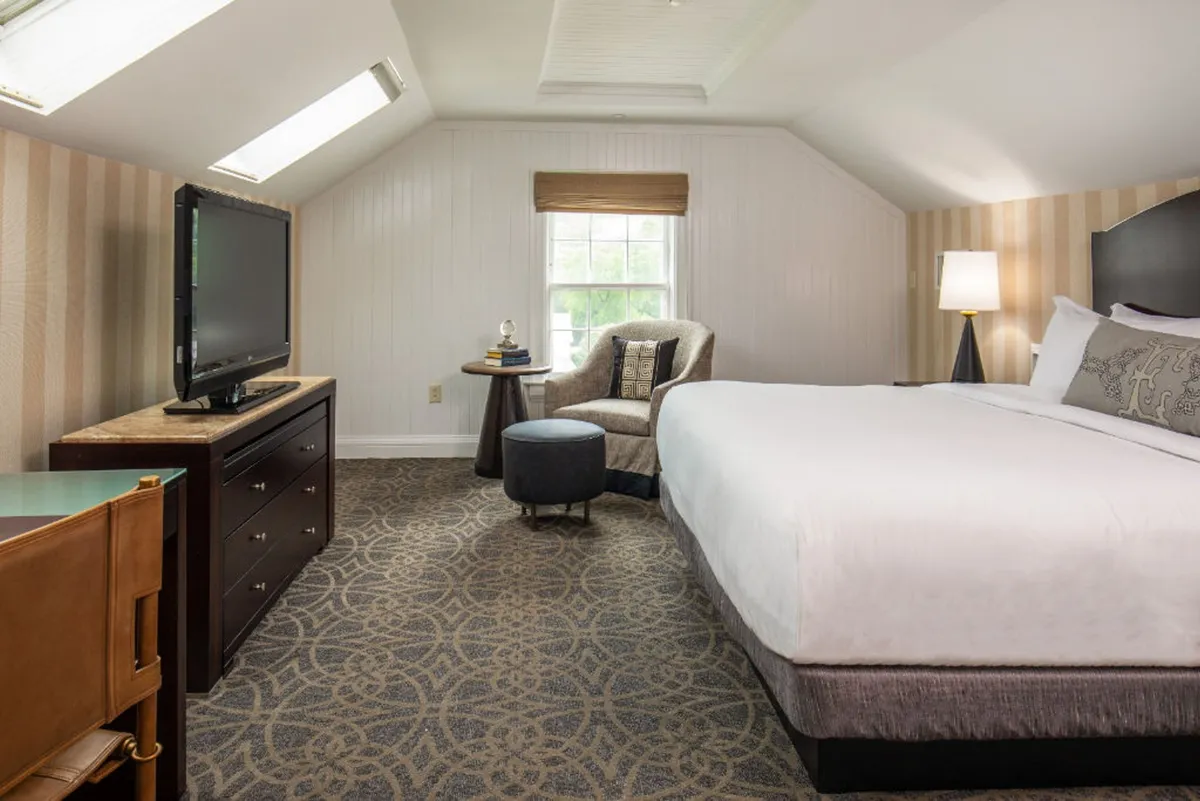 The image showcases a serene, sunlit guest room at The Equinox Golf Resort & Spa, featuring a plush king-sized bed, a cozy armchair for reading, a sleek television for unwinding, and a large window that fills the space with natural light. The interiors blend classic New England charm with modern comfort, creating a restful retreat.