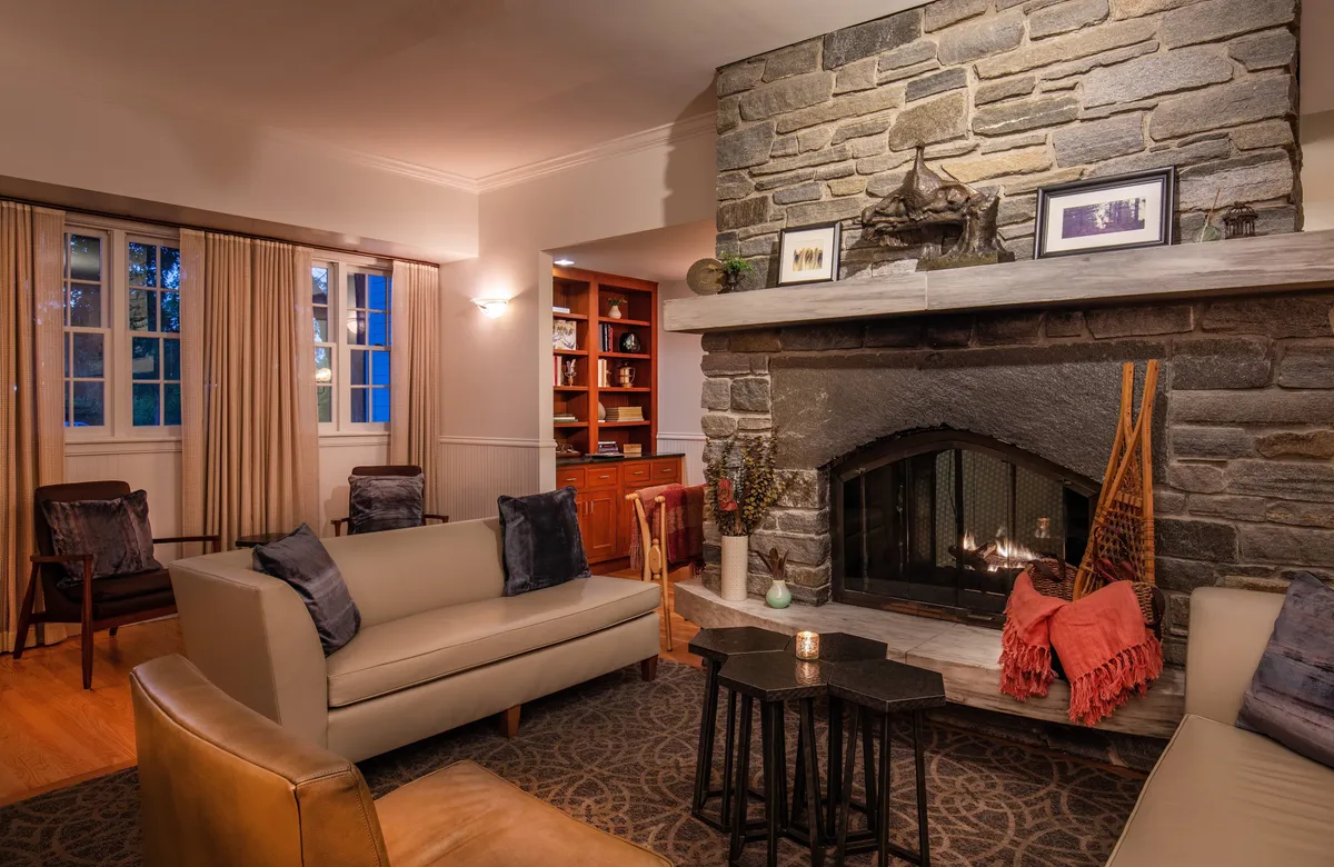 A cozy interior space highlighted by a stone fireplace, built-in bookshelf, and inviting sofa. The warm, glowing fire and traditional New England-inspired accents create an immediate sense of comfort and relaxation. Subtle holiday decorations and classic furnishings evoke both history and hospitality.