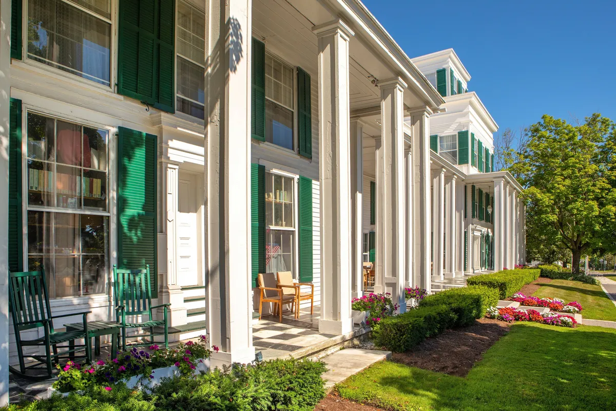 A classic New England porch with stately white columns and green shutters, lined with comfortable rocking chairs and bordered by vibrant flower beds. The mood is welcoming and tranquil, suggesting a place to unwind and soak in village life, perhaps before or after an evening out.
