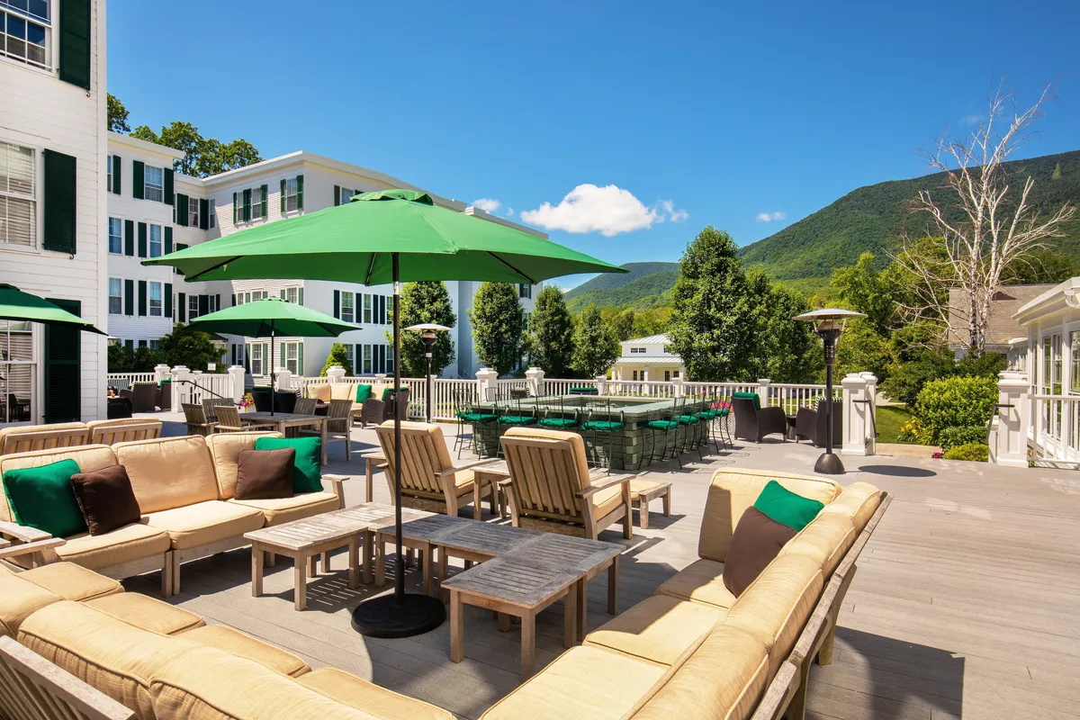 This image showcases an inviting outdoor seating area set under green umbrellas, with guests relaxing and socializing against a breathtaking mountain backdrop. The scene exudes a leisurely, tranquil atmosphere ideal for outdoor dining or enjoying cocktails as the sun sets.