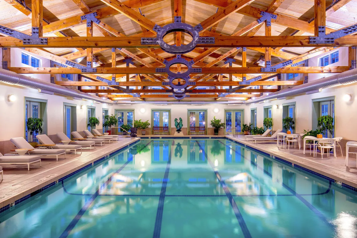 A sophisticated interior featuring an inviting indoor swimming pool accented by wooden beams, contemporary art sculptures, and plush lounge chairs. The overall mood is serene and luxurious, hinting at the exclusive feel of the suite's amenities.