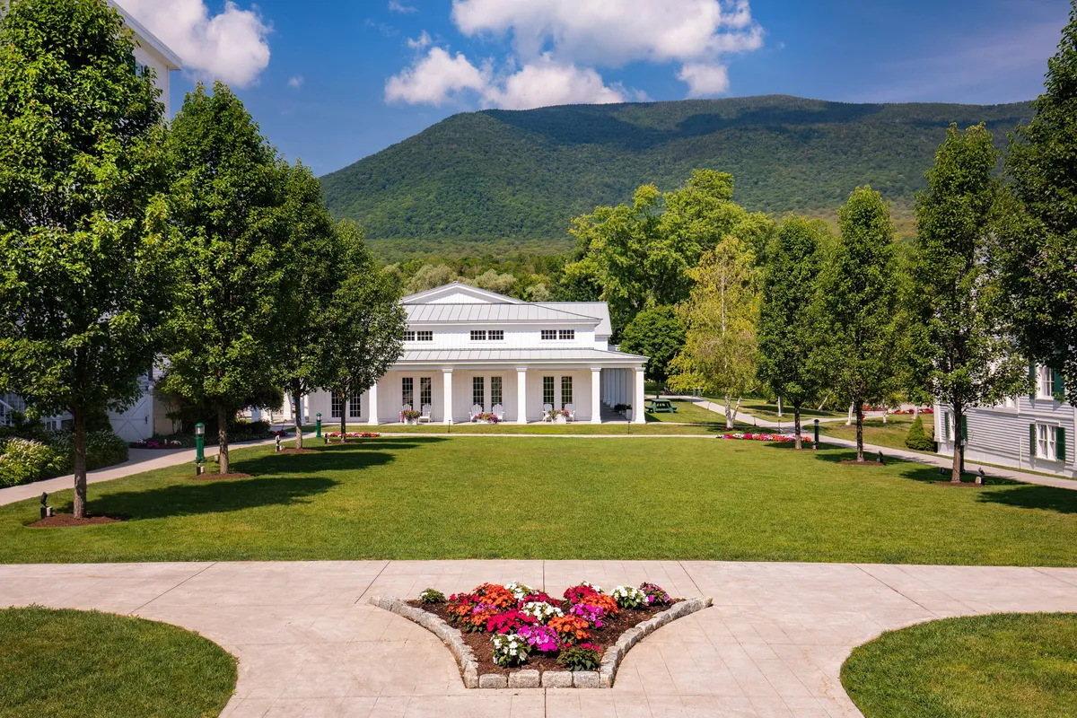 A vibrant exterior shot of manicured lawns, colorful flower beds, and a crisp white building nestled against a majestic mountain backdrop. The scene suggests plenty of open space for outdoor gatherings, relaxation, and wellness activities such as yoga or seasonal events.