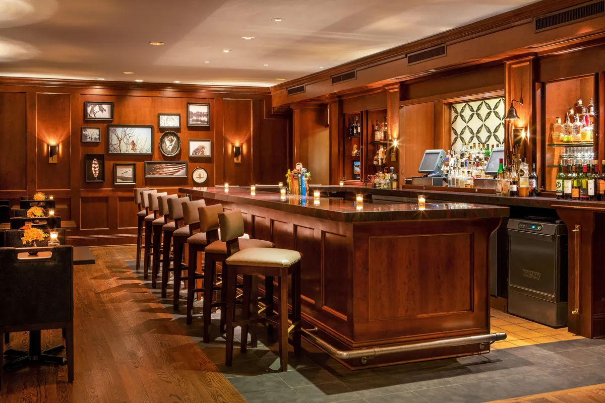 This inviting interior scene centers on a sleek bar counter with several comfortable stools lined up, stylish wall art, and a curated selection of bottled drinks. The setup encourages socializing and unwinding, embodying the loft's spirit of connection and memorable moments with loved ones.