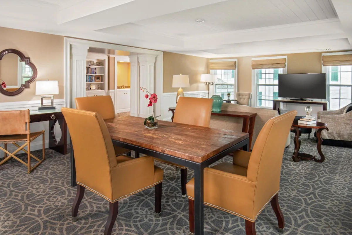 This interior image features a cozy, open-concept dining area with a prominent wooden dining table surrounded by leather chairs. A decorative vase with fresh flowers graces the table, and a TV unit anchors the background, reinforcing the space as a central gathering place for meals, games, and movie nights. The scene is warmly lit, evoking a sense of comfort and invitation.