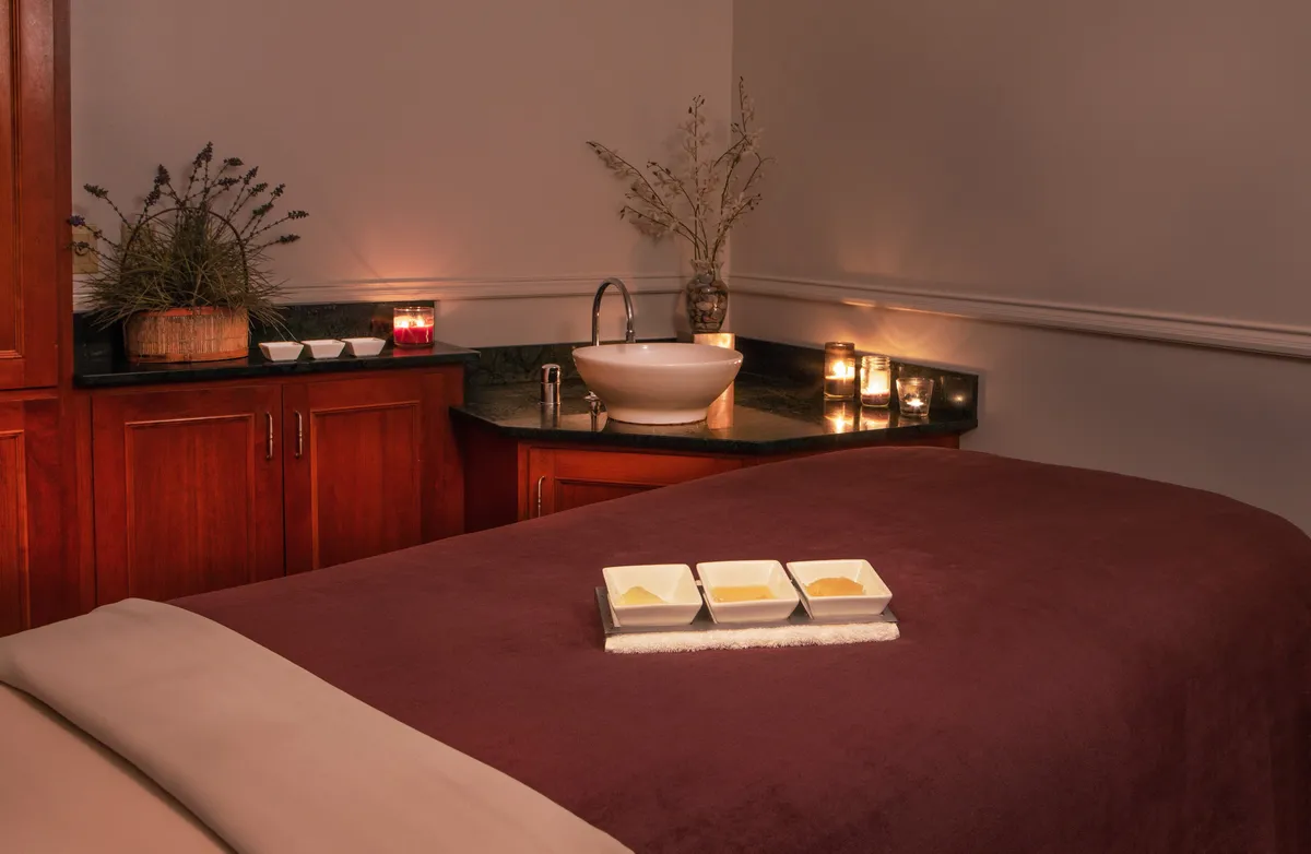 This interior image features a serene massage table set up with a bowl of oils, candles, and a nearby sink. The decor is tranquil and spa-inspired, evoking a strong sense of relaxation and personal pampering.