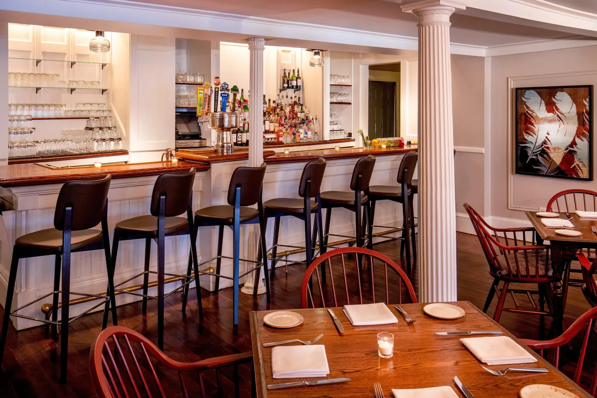A sophisticated interior showing a bar counter with stools, a dining table, and art adorning the wall. The space blends classic and modern elements, inviting guests to gather, socialize, or enjoy quiet moments. Warm colors and stylish touches enhance its welcoming vibe.